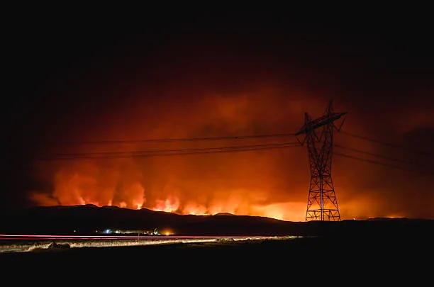 Transmission tower and power lines silhouetted against wildfire glow at night